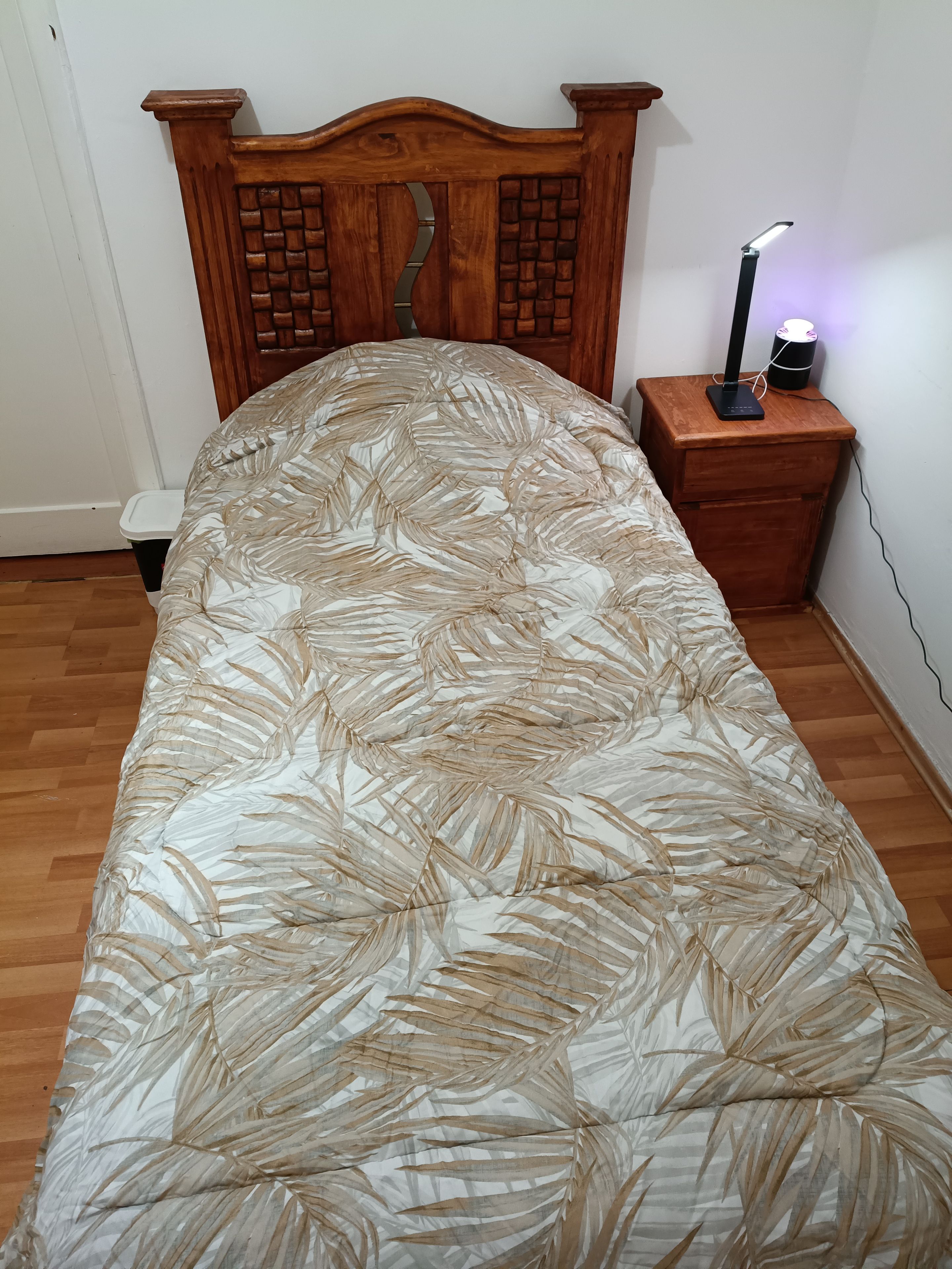Un lit avec une couvre-lit à motifs de feuilles, une tête de lit en bois, une table de chevet avec une lampe et une prise de courant.