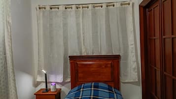 Un dormitorio con cabecera de madera, cama con colcha a cuadros azul y blanco, cortinas blancas, lámpara de noche y mesita de noche de madera.