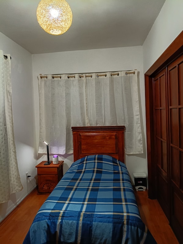 Une chambre à coucher avec une tête de lit en bois, un lit recouvert d’une couverture à carreaux bleus et blancs, des rideaux blancs, une lampe de chevet et une petite table de chevet en bois.