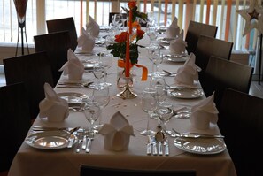 Una larga mesa de comedor dispuesta con servilletas blancas, cubiertos y copas de vino, con un centro de mesa con una rosa y una vela.