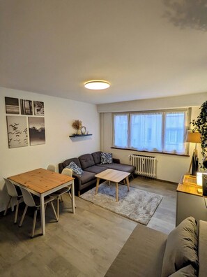 A modern living room with a sofa, a dining table, and chairs. There is a radiator, a window with blinds, and a ceiling light. - Appartement cosy proche du centre de Strasbourg (Strasbourg)