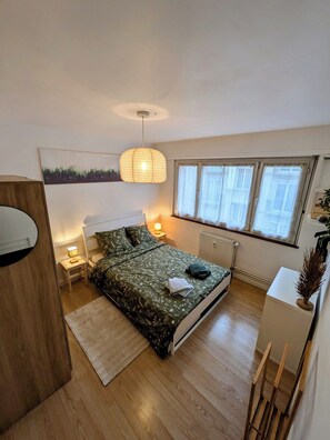A bedroom with a bed, bedside table, lamp, window, radiator, and a small desk with a plant. - Appartement cosy proche du centre de Strasbourg (Strasbourg)