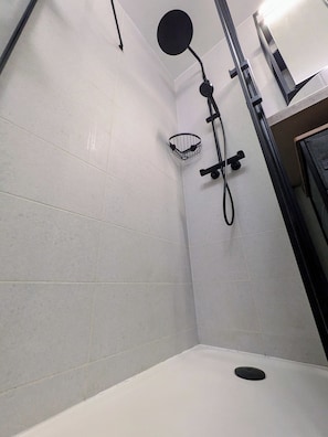 Rincón de un baño con paredes de azulejos blancos, una ducha, un cabezal de ducha y una pequeña cesta metálica.
