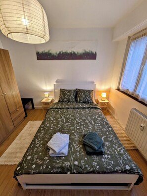 A bedroom with a bed, two bedside tables, a nightstand, a radiator, a window with curtains, and a wooden wardrobe. - Appartement cosy proche du centre de Strasbourg (Strasbourg)