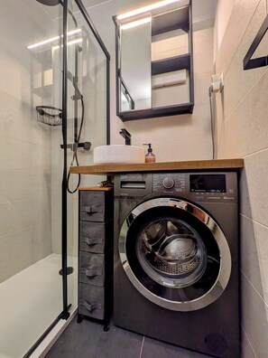 Une salle de bain compacte équipée d’un lave-linge intégré, d’une cabine de douche en verre et d’un miroir au-dessus du lavabo.
