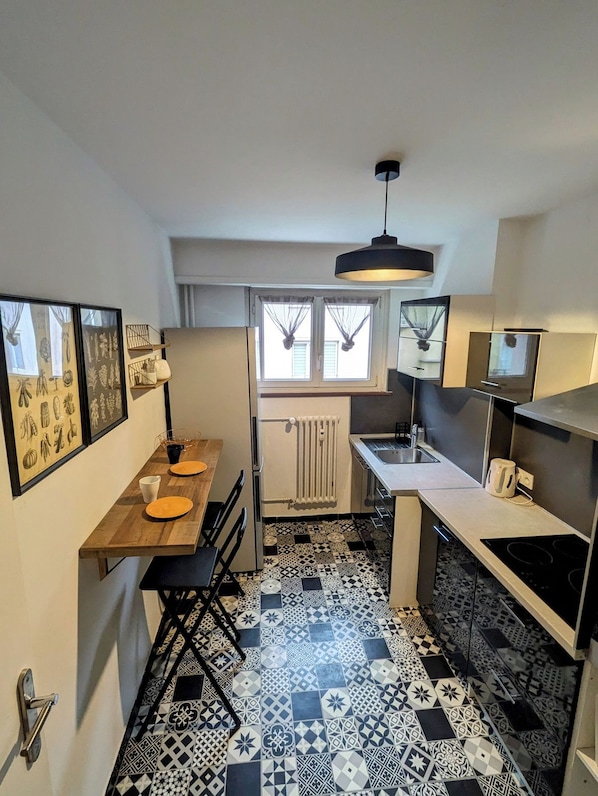 A compact kitchen with a dining area, a radiator, and a window with curtains. - Appartement cosy proche du centre de Strasbourg (Strasbourg)