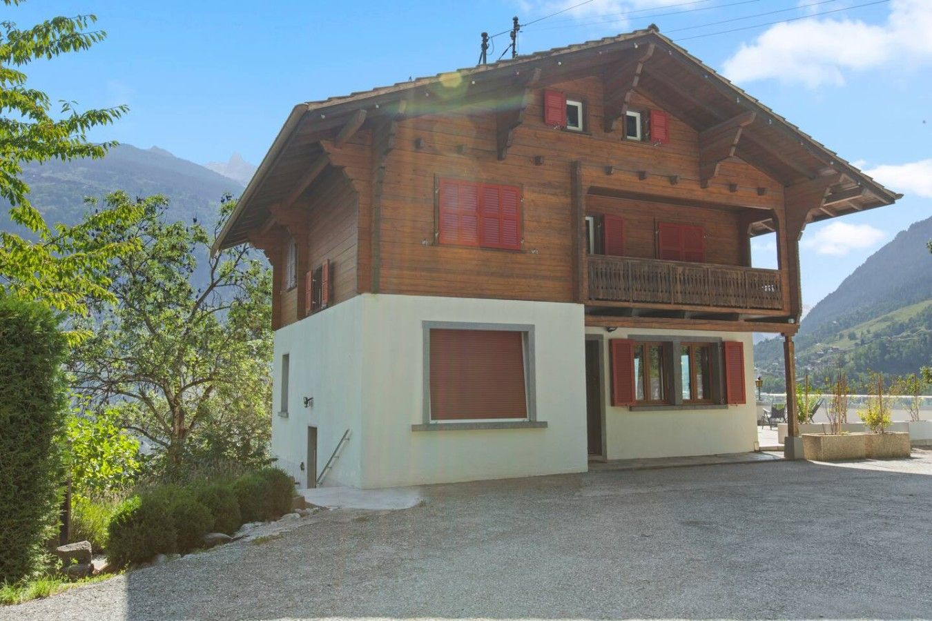A two-story wooden chalet with a balcony, set against a mountainous backdrop.