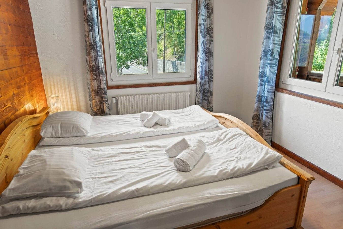 A double bed with white linens and towels, a wooden headboard, a window with curtains, and a radiator.