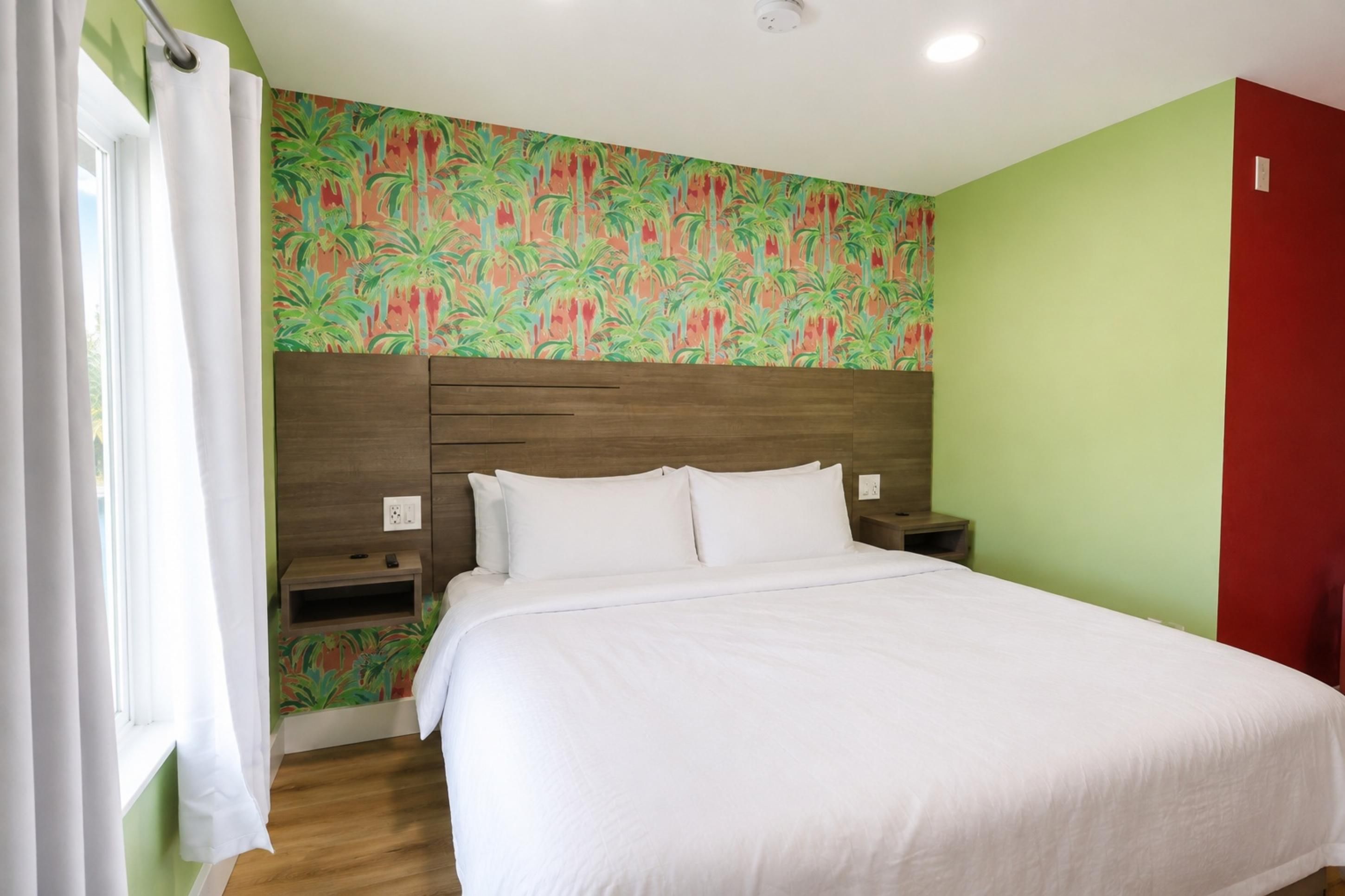 Habitación de hotel con una cama, mesitas de noche, una ventana con cortinas y un papel tapiz tropical en la pared.