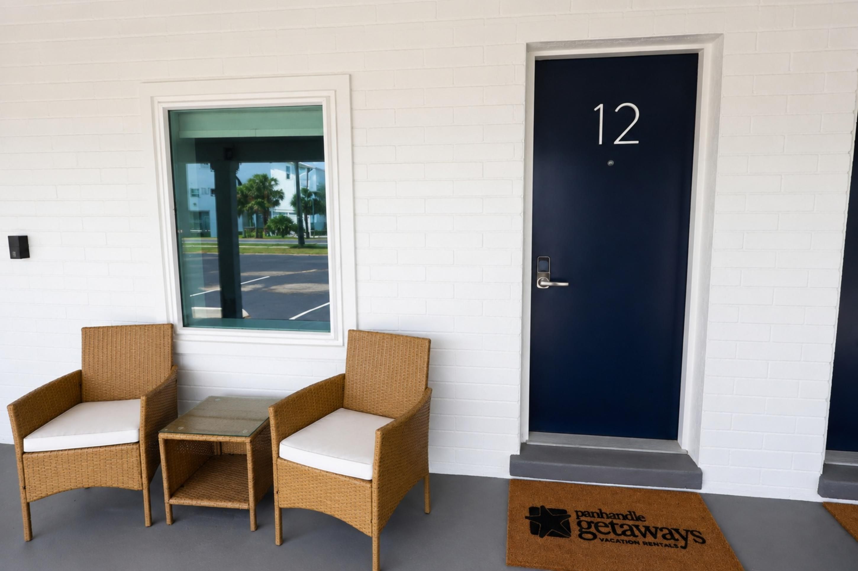 A hotel entrance with a dark blue door marked with the number 12, two wicker chairs with white cushions, and a view of a parking lot through a glass door.