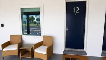 A hotel entrance with a dark blue door marked with the number 12, two wicker chairs with white cushions, and a view of a parking lot through a glass door.