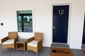 En hotellentrél med en mörkblå dörr som är numrerad med 12, två flätade fåtöljer med vita kuddar och en utsikt över en parkeringsplats genom en glasdörr.