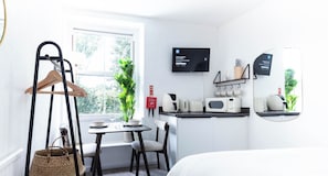 A compact room with a bed, a small table with a chair, a hanging clothes rack, a microwave, a small shelf with cups, and a fire blanket.