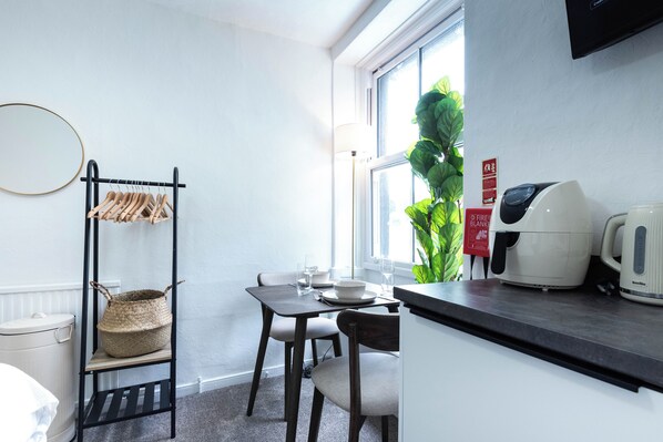 Une cuisine compacte comprenant une table à manger, des chaises, une bouilloire et une fenêtre donnant sur une plante à l’extérieur.