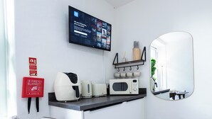 Cozinha compacta equipada com micro-ondas, torradeira e chaleira, manta anti-fogo, TV exibindo opções de filmes e um espelho.