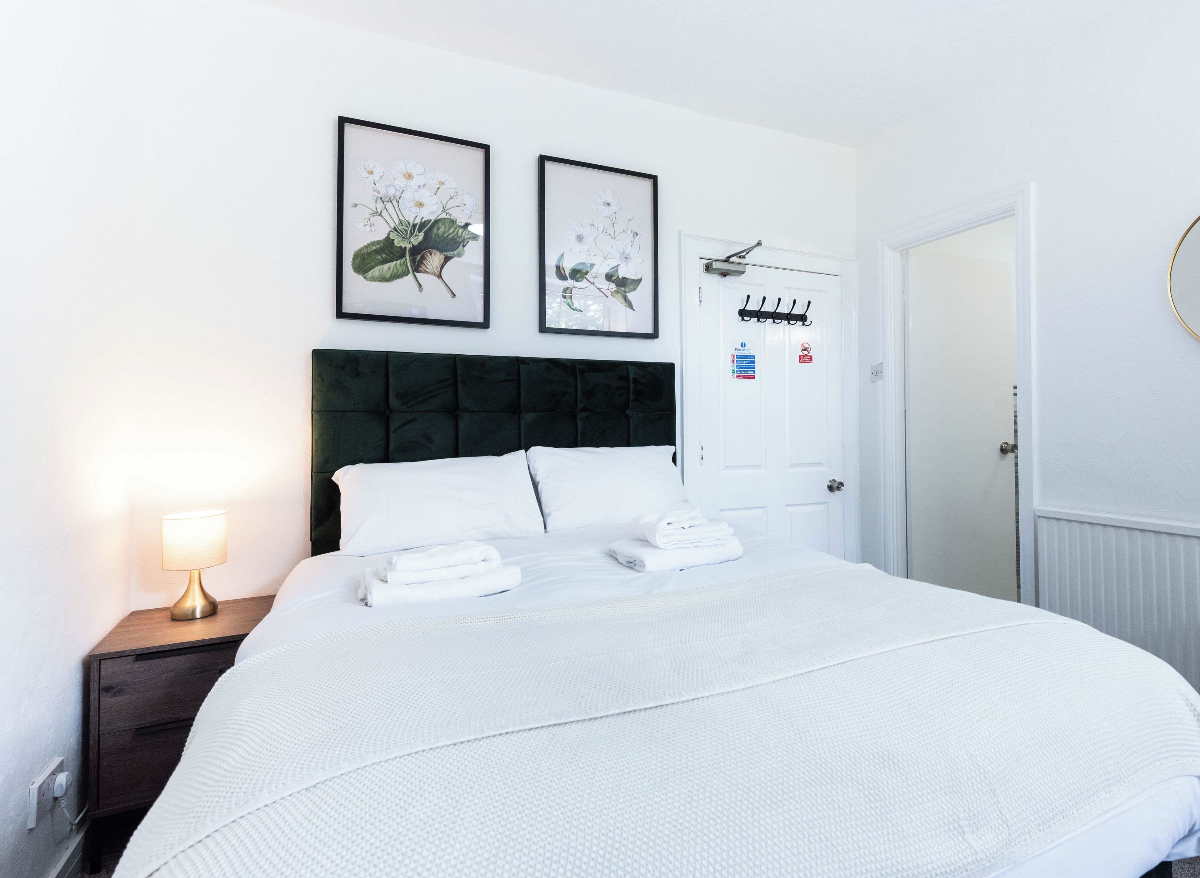 A hotel room with a bed, a nightstand with a lamp, and two framed botanical prints on the wall.