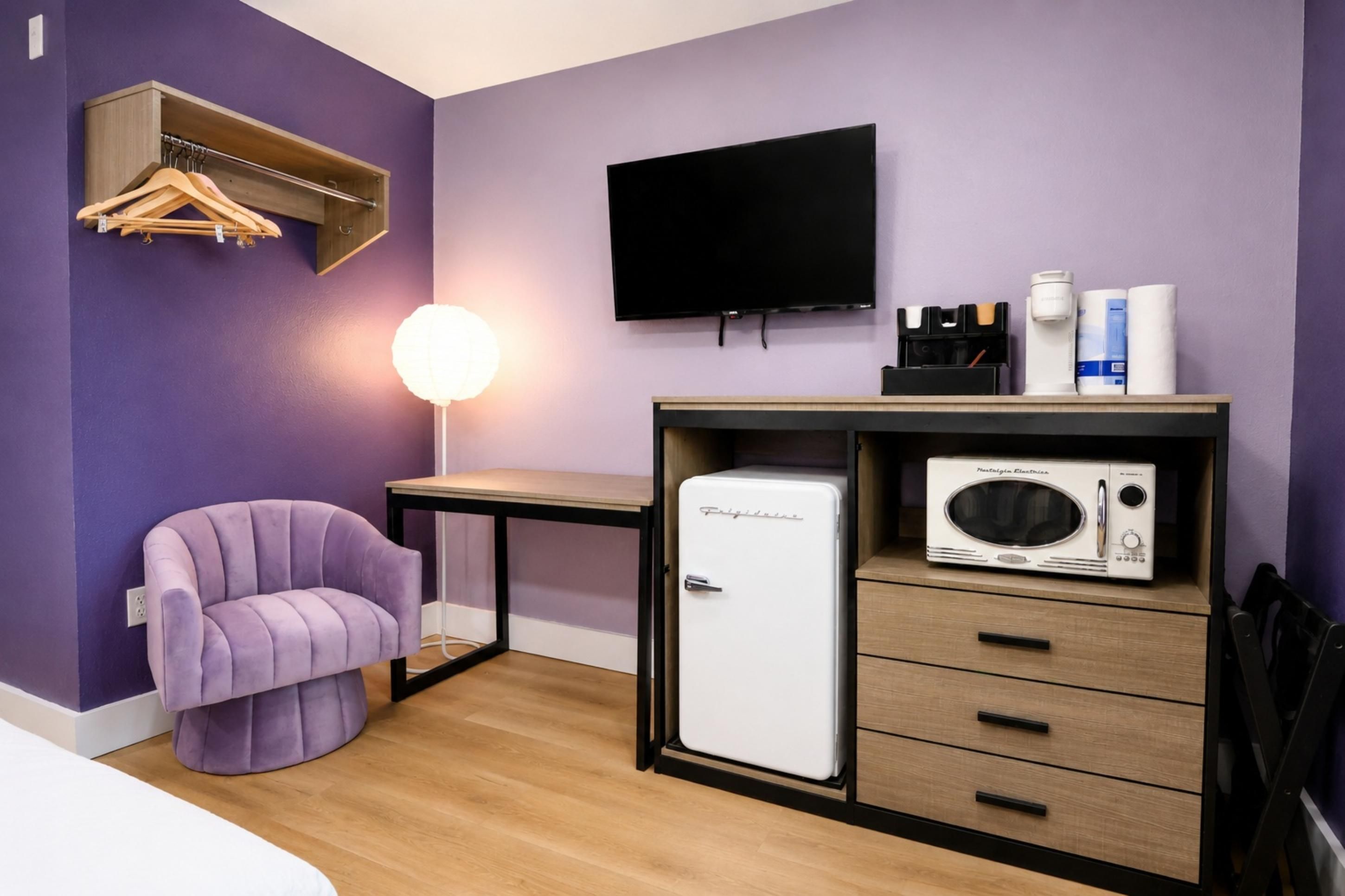 A compact room with a purple wall, a single bed, a small desk with a flat-screen TV, a white refrigerator, and a chair.