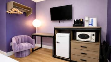 A compact room with a purple wall, a single bed, a small desk with a flat-screen TV, a white refrigerator, and a chair.
