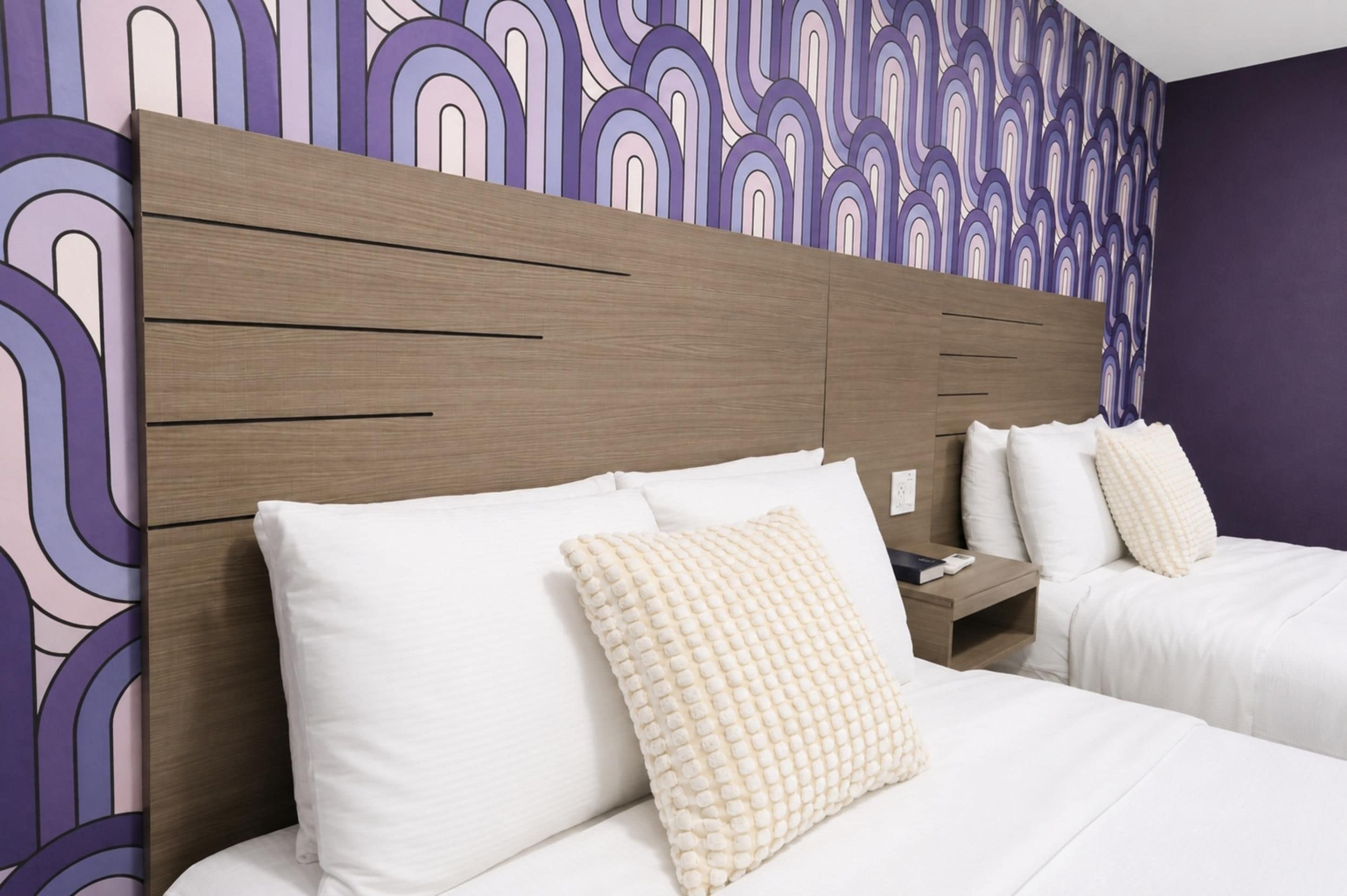 A hotel room with a modern headboard, two beds with white linens, and a patterned wallpaper.