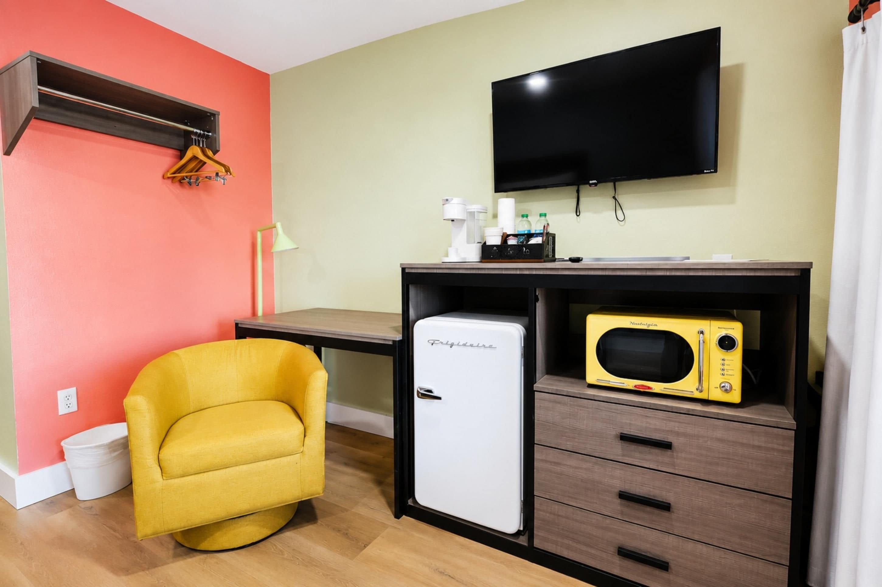 A compact room with a yellow chair, a small refrigerator, a microwave, a flat-screen TV, and a desk with a lamp and a coffee maker.