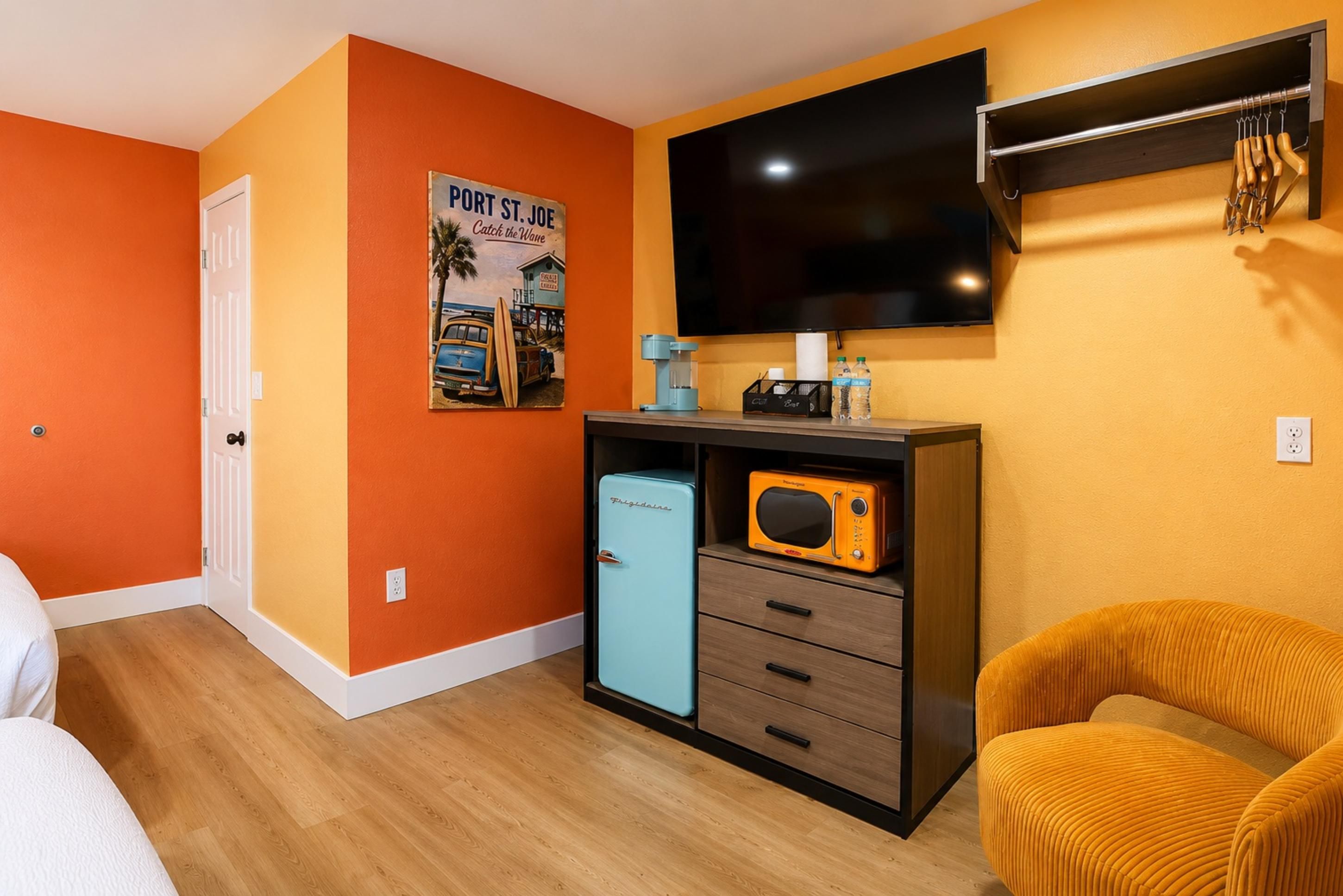 Habitación con paredes anaranjadas, un televisor montado en la pared, un refrigerador pequeño, un microondas, una cafetera y una silla.