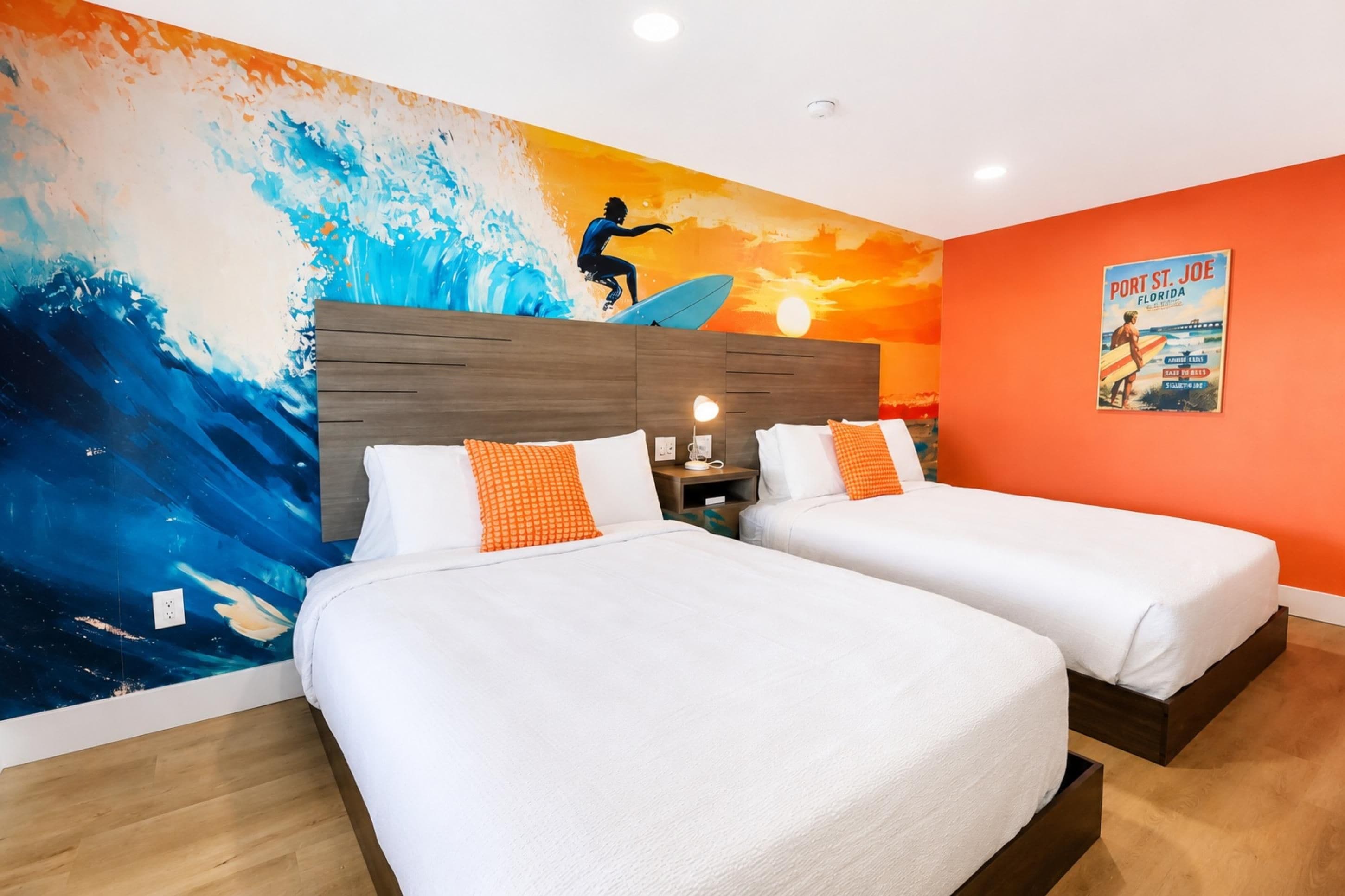 A hotel room with two beds, a mural of a surfer on a wave, and a poster of Port St. Joe, Florida.