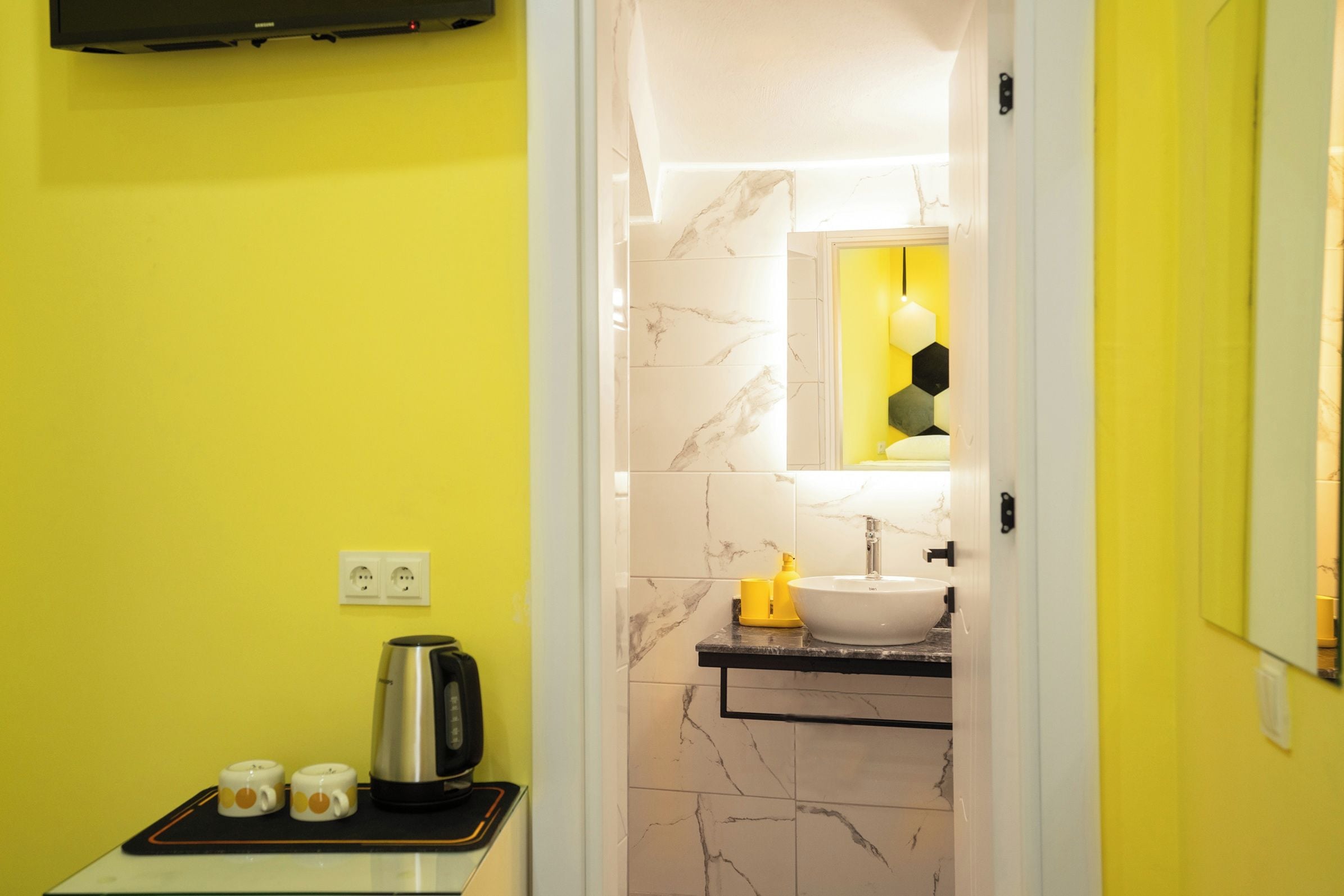 Een moderne badkamer met een gele wand, een wand betegeld met marmer, een wastafel met spiegel, en een waterkoker op een klein tafeltje.