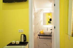 Un baño moderno con pared amarilla, pared revestida de mármol, lavamanos con espejo y una tetera sobre una mesita.