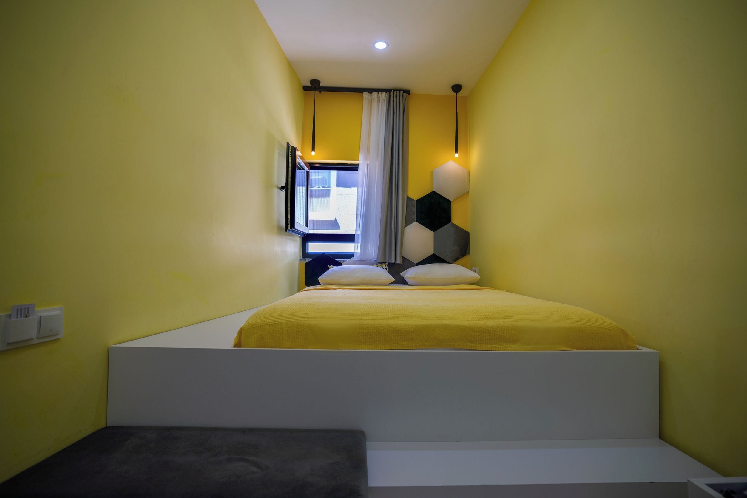 Quarto de hotel estreito com cama, um banquinho e janela com cortinas.