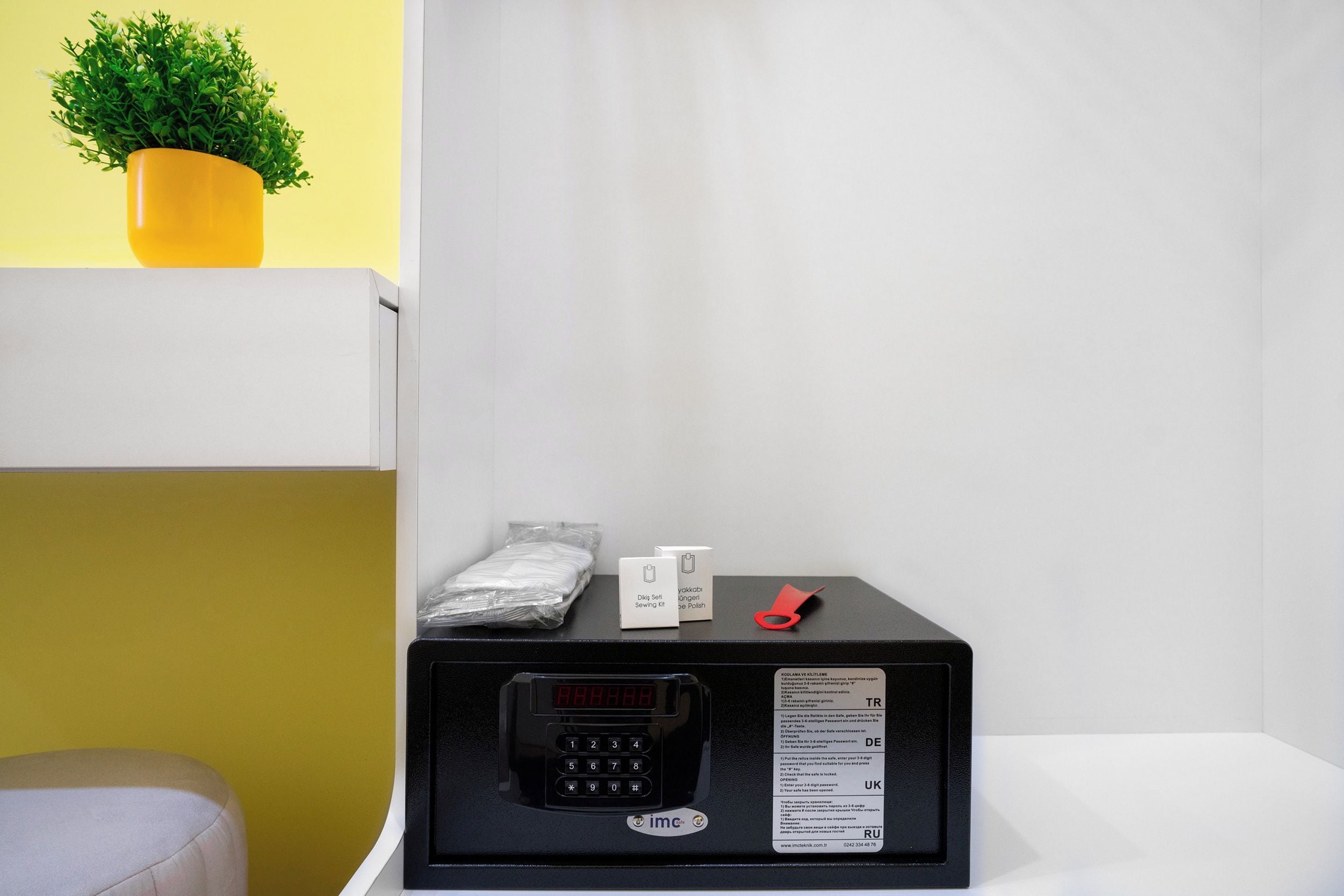 A black microwave on a white table with a small potted plant, a red object, and a sealed package.