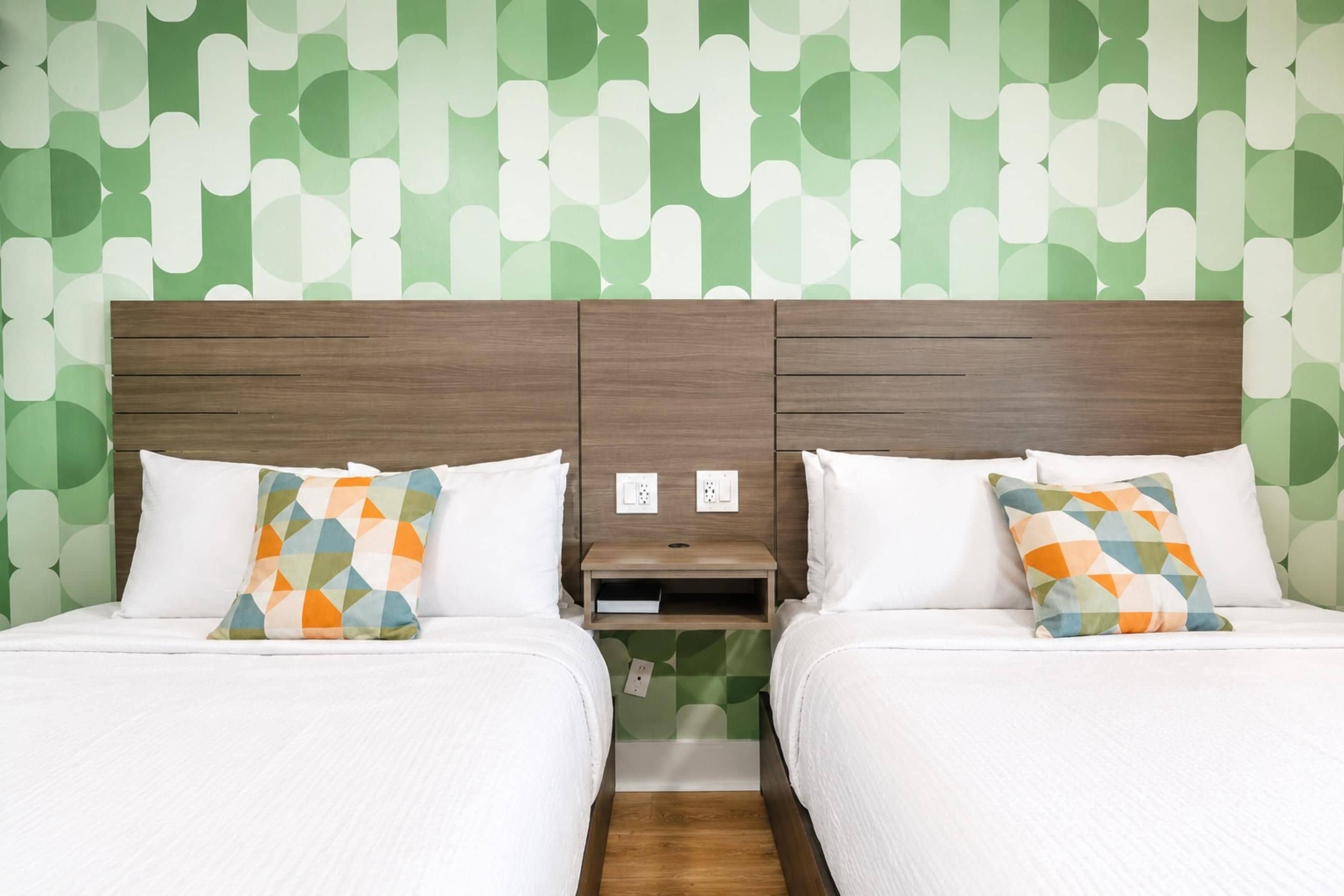 A hotel room with two beds, each with a patterned pillow, a wooden headboard, and a nightstand with a power outlet.