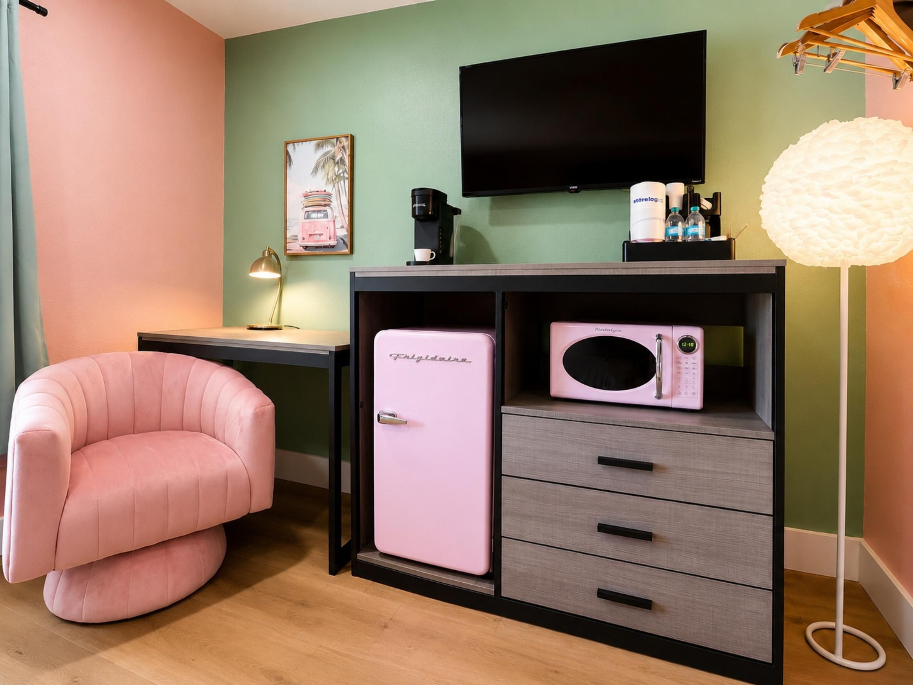 A room with a pink chair, a small fridge, a desk with a lamp, a television, and a white lamp.
