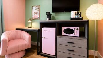 A room with a pink chair, a small fridge, a desk with a lamp, a television, and a white lamp.