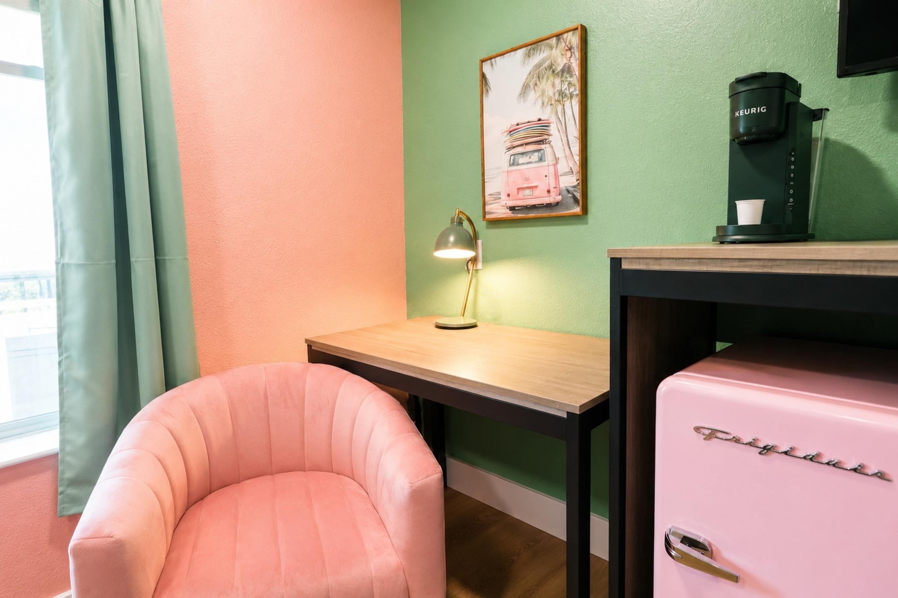 Una habitación con una silla rosa, una pequeña mesa de madera, una lámpara, una cafetera y un cuadro en la pared.