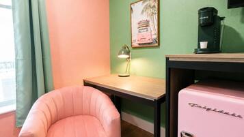 Una habitación con una silla rosa, una pequeña mesa de madera, una lámpara, una cafetera y un cuadro en la pared.