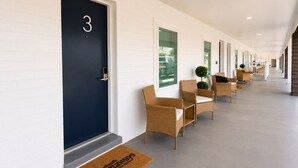 Un couloir d'hôtel avec des portes numérotées, des chaises en osier et un paillasson avec du texte.