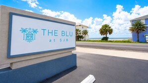 Placa de 'The Blu at SJB', com céu claro e palmeiras ao fundo.