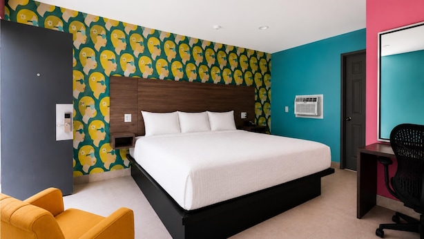 Quarto de hotel moderno com uma cama grande, uma escrivaninha, uma cadeira e uma parede com padrão repetido.