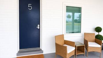 Una puerta azul oscuro con el número 5, una pequeña mesa con sillas y una ventana con vistas a palmeras.
