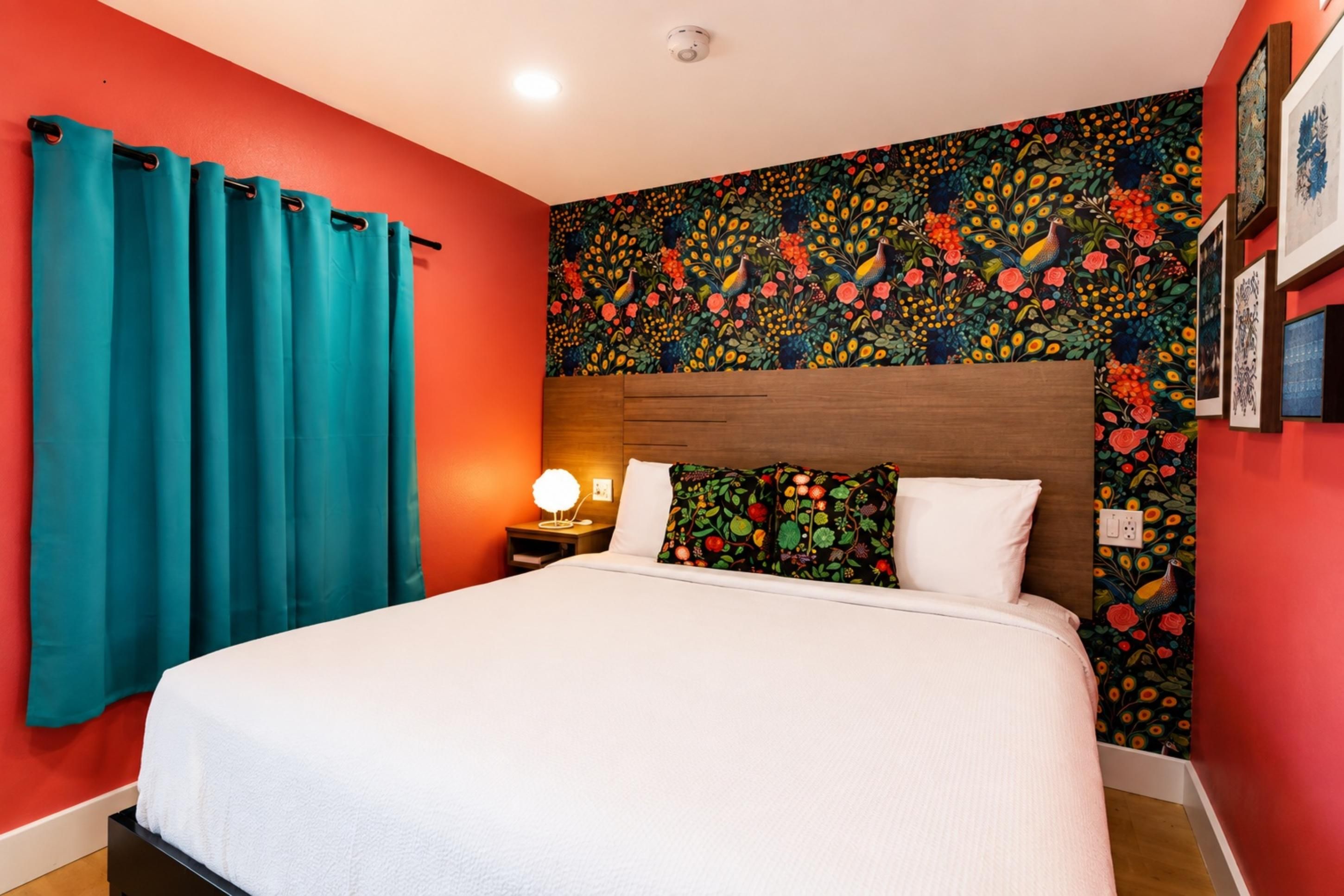 Una habitación de hotel con cama, cortinas azul turquesa, un ventanal con estampado floral y una mesita de noche con lámpara.