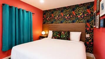 Una habitación de hotel con cama, cortinas azul turquesa, un ventanal con estampado floral y una mesita de noche con lámpara.