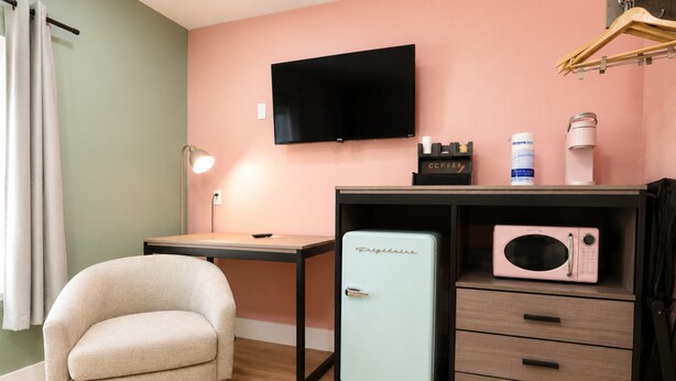 Quarto compacto com cama, escrivaninha, cadeira, televisão, micro-ondas e um pequeno frigorífico.