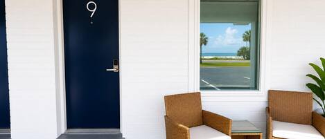 Habitación con una puerta azul oscuro marcada con el número 9, sillas de mimbre con cojines blancos, una mesita y vista a una playa a través de una ventana.