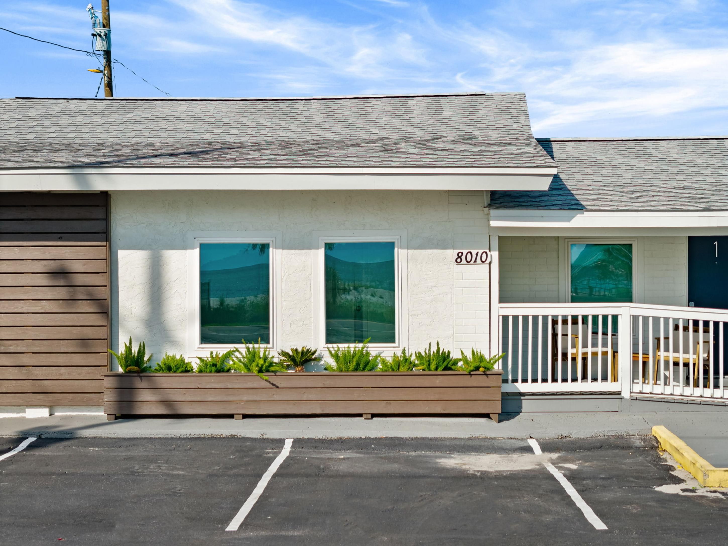 A single-story building with a white exterior, numbered 8010, featuring a porch with a white railing and a small seating area.