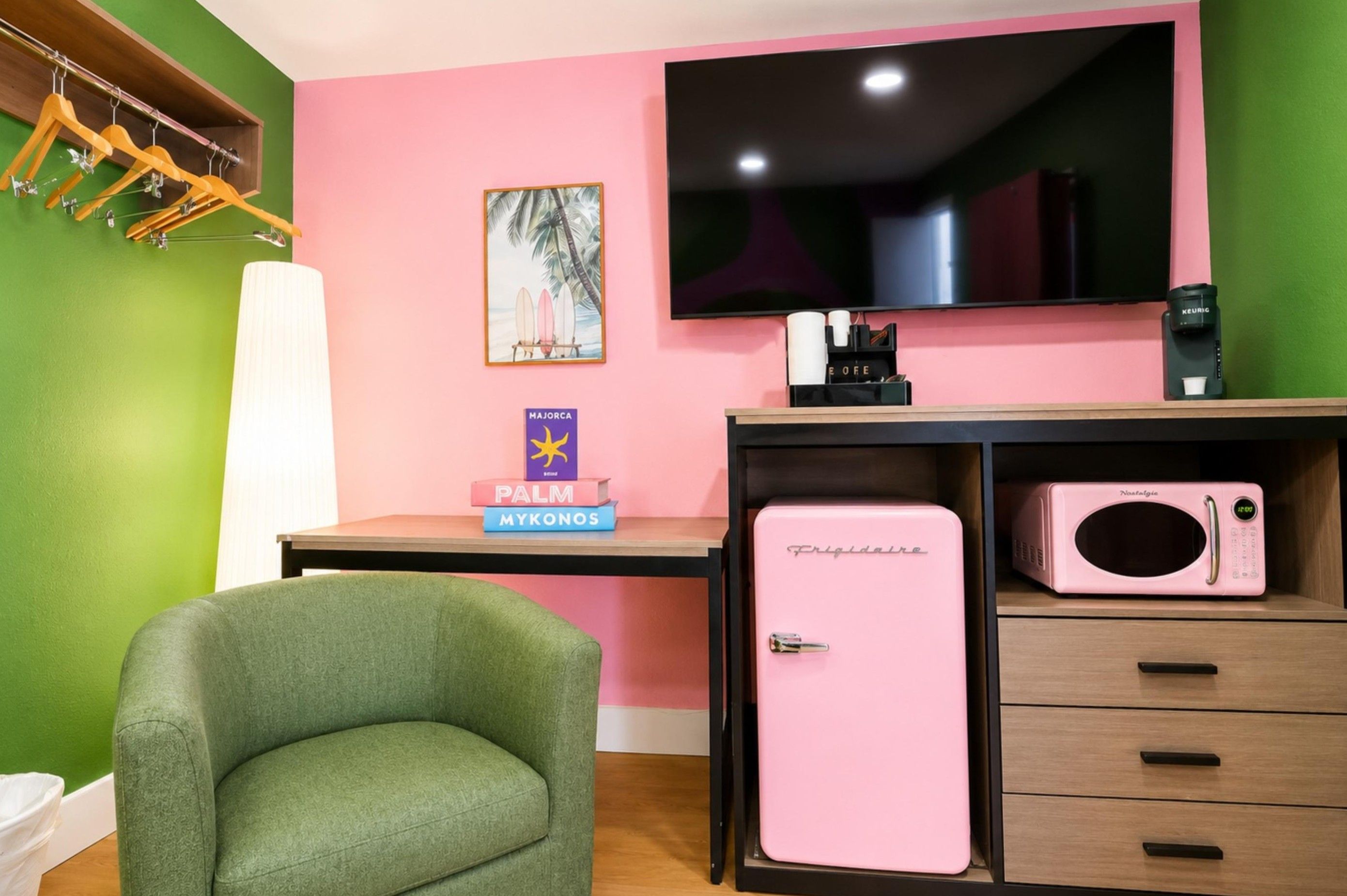 A room with a green wall, a pink sofa, a small pink refrigerator, a coffee machine, and a TV on the wall.