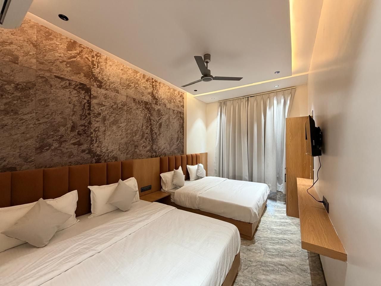 Quarto de hotel com duas camas, um ventilador de teto, cabeceira de madeira e uma parede grande com textura.