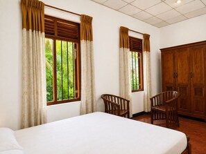 A hotel room with two single beds, a wooden wardrobe, two wooden chairs, and windows with curtains. - Piyasewana Spice Garden Villa (Banduragoda)
