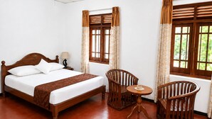 A hotel room with a wooden bed, two chairs, a small table, and windows with curtains. - Piyasewana Spice Garden Villa (Banduragoda)