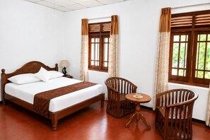 A hotel room with a wooden bed, two chairs, a small table, and windows with curtains. - Piyasewana Spice Garden Villa (Banduragoda)