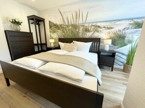 A bedroom with a large bed, a dresser, a nightstand, and a view of the beach through the window. - Holiday Apartment With 1 Bedroom (Cuxhaven)